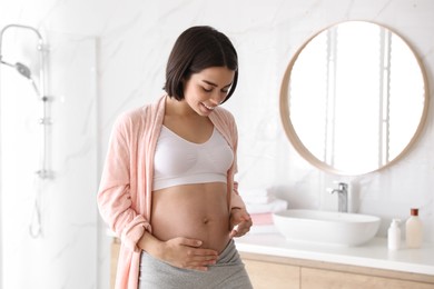 Photo of Young pregnant woman applying cosmetic product on belly in bathroom