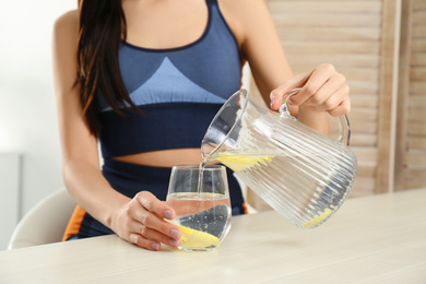 Sportive woman pouring lemon water into glass from jug at table indoors, closeup