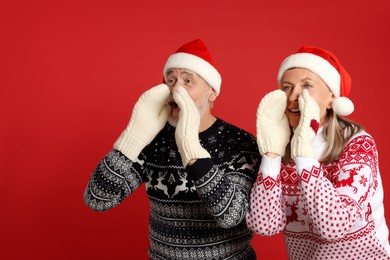 Photo of Senior couple in Christmas sweaters, Santa hats and knitted mittens shouting something on red background