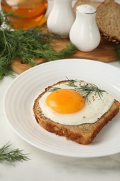 Plate with tasty fried egg, slice of bread and dill on white marble table, closeup