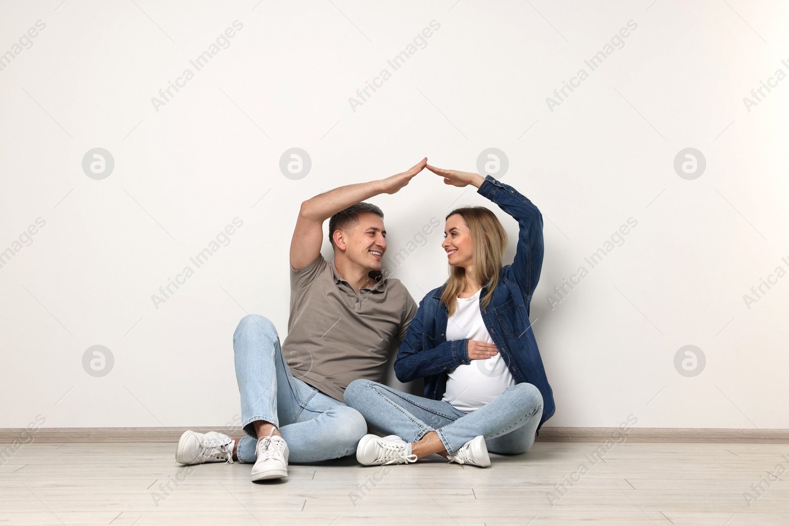 Photo of Young family housing concept. Pregnant woman with her husband forming roof with their hands while sitting on floor indoors