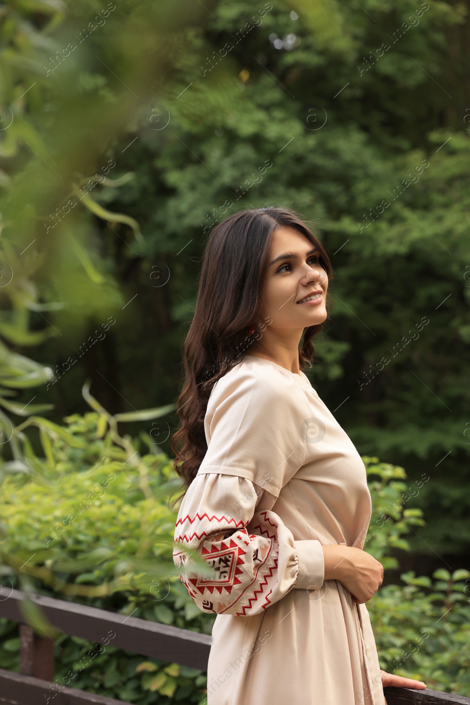 Photo of Beautiful woman wearing embroidered dress near wooden railing in countryside, space for text. Ukrainian national clothes