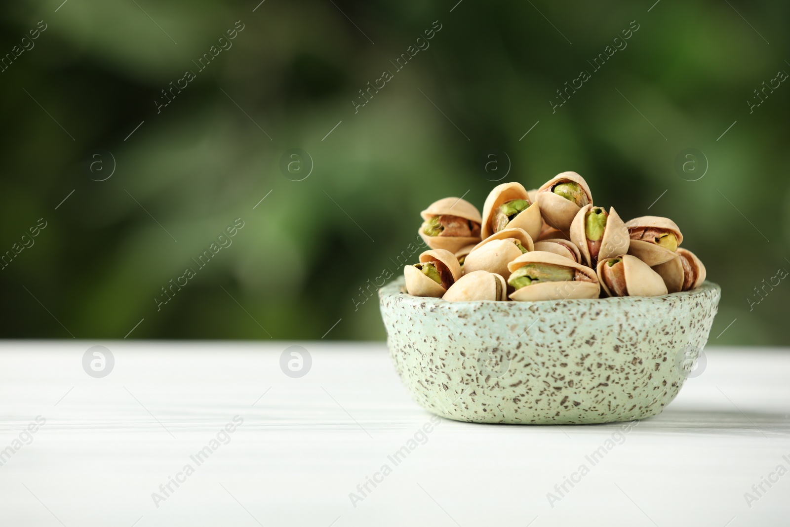 Photo of Tasty pistachios in bowl on white table against blurred background. Space for text