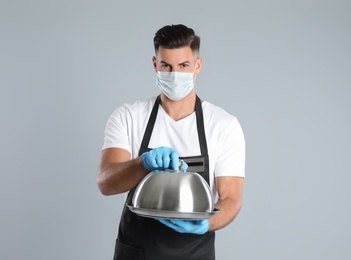 Photo of Waiter in medical face mask holding tray with lid on light grey background