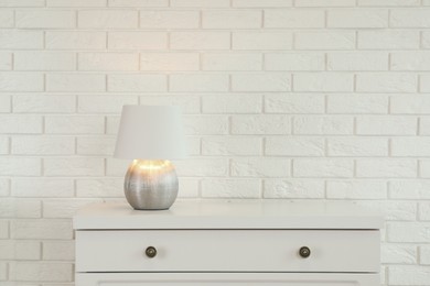 Modern chest of drawers with lamp near white brick wall