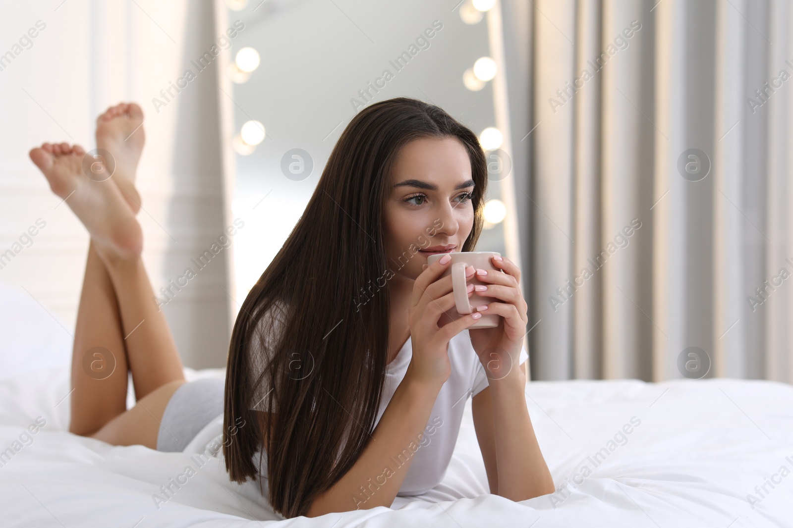 Photo of Young woman in white t-shirt and elegant underwear with cup of drink on bed indoors