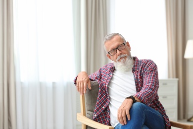 Photo of Portrait of handsome mature man with glasses sitting in armchair indoors. Space for text