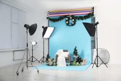 Photo of Beautiful Christmas themed photo zone with professional equipment, stylish armchair, trees and gift boxes in studio