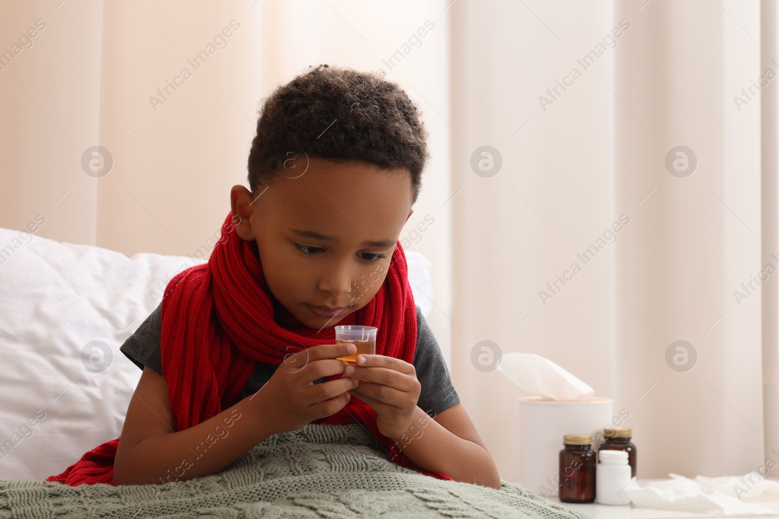 Photo of African-American boy taking cough syrup on bed at home, space for text. Cold medicine