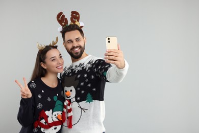Happy young couple in Christmas sweaters and reindeer headbands taking selfie on grey background. Space for text