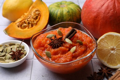 Photo of Bowl of delicious pumpkin jam and ingredients on tiled surface, closeup