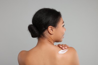 Young woman applying body cream onto back on grey background