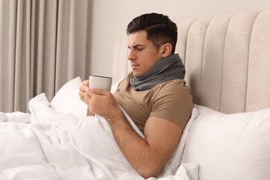 Photo of Sick man with cup of hot drink and scarf in bed at home