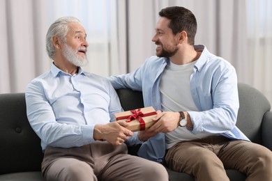 Photo of Son giving gift box to his dad on sofa at home