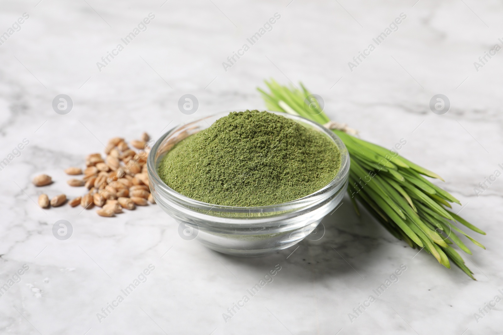 Photo of Wheat grass powder in glass bowl, seeds and fresh sprouts on white marble table, closeup