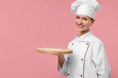 Photo of Happy woman chef in uniform holding empty wooden board on pink background, space for text