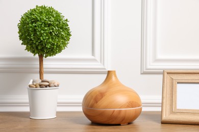 Green artificial plant in pot, frame and air humidifier on wooden table near white wall