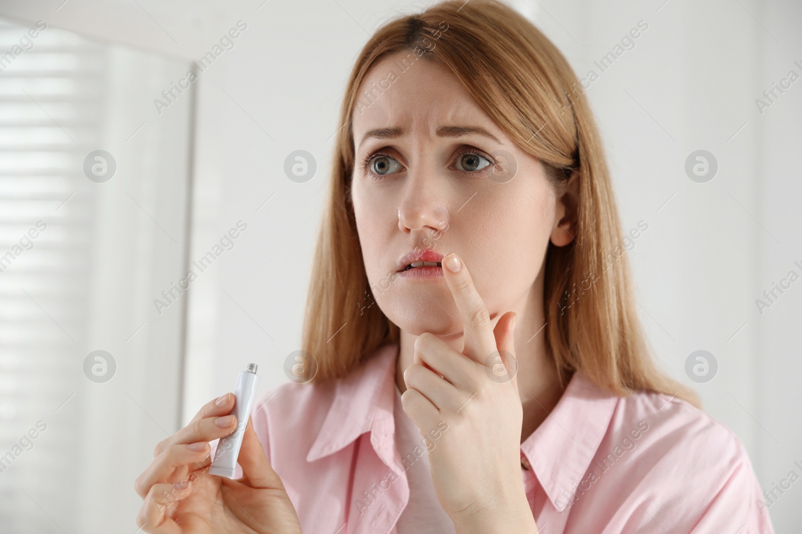 Photo of Upset woman with herpes applying cream on lips indoors