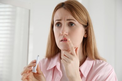 Upset woman with herpes applying cream on lips indoors