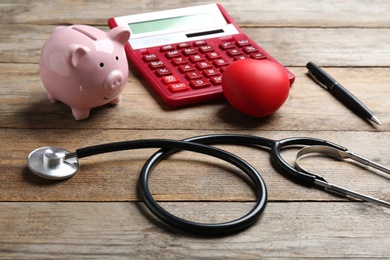 Photo of Stethoscope, red heart and calculator with piggybank on wooden surface. Health insurance concept