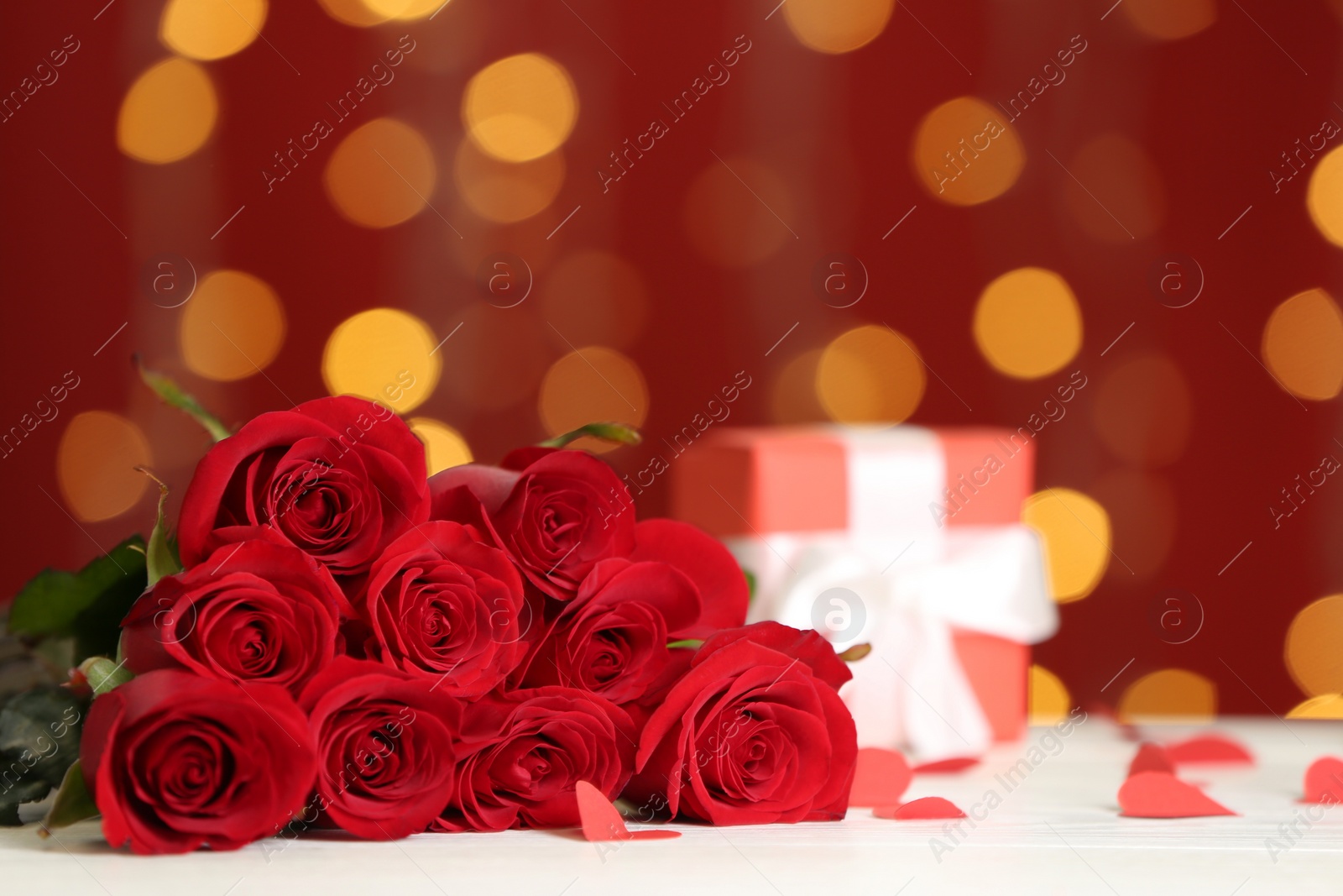 Photo of Beautiful red roses and gift box on white table against blurred lights, space for text. St. Valentine's day celebration