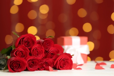 Photo of Beautiful red roses and gift box on white table against blurred lights, space for text. St. Valentine's day celebration