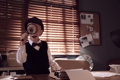 Photo of Cute little detective with magnifying glass at table in office. Space for text