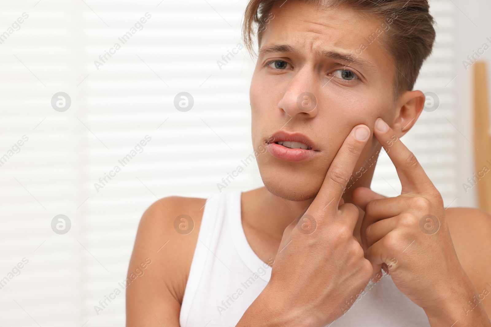 Photo of Upset young man popping pimple on his face indoors. Acne problem