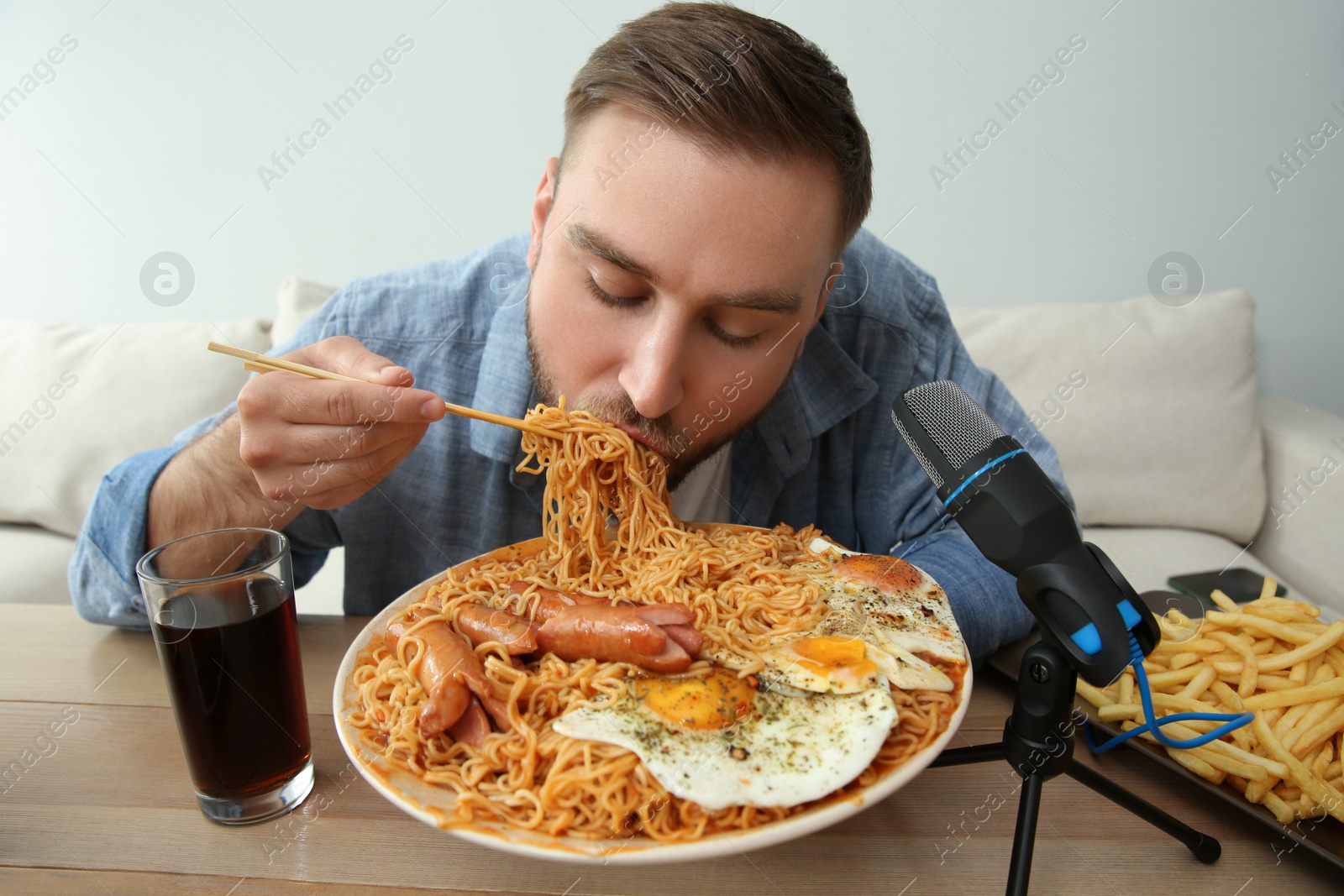 Photo of Food blogger eating in front of microphone at table against light background. Mukbang vlog