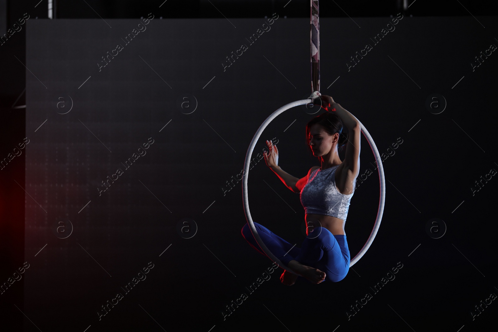 Photo of Young woman performing acrobatic element on aerial ring against dark background. Space for text