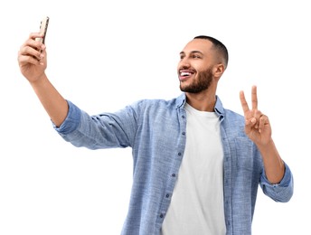 Photo of Smiling young man taking selfie with smartphone and showing peace sign on white background