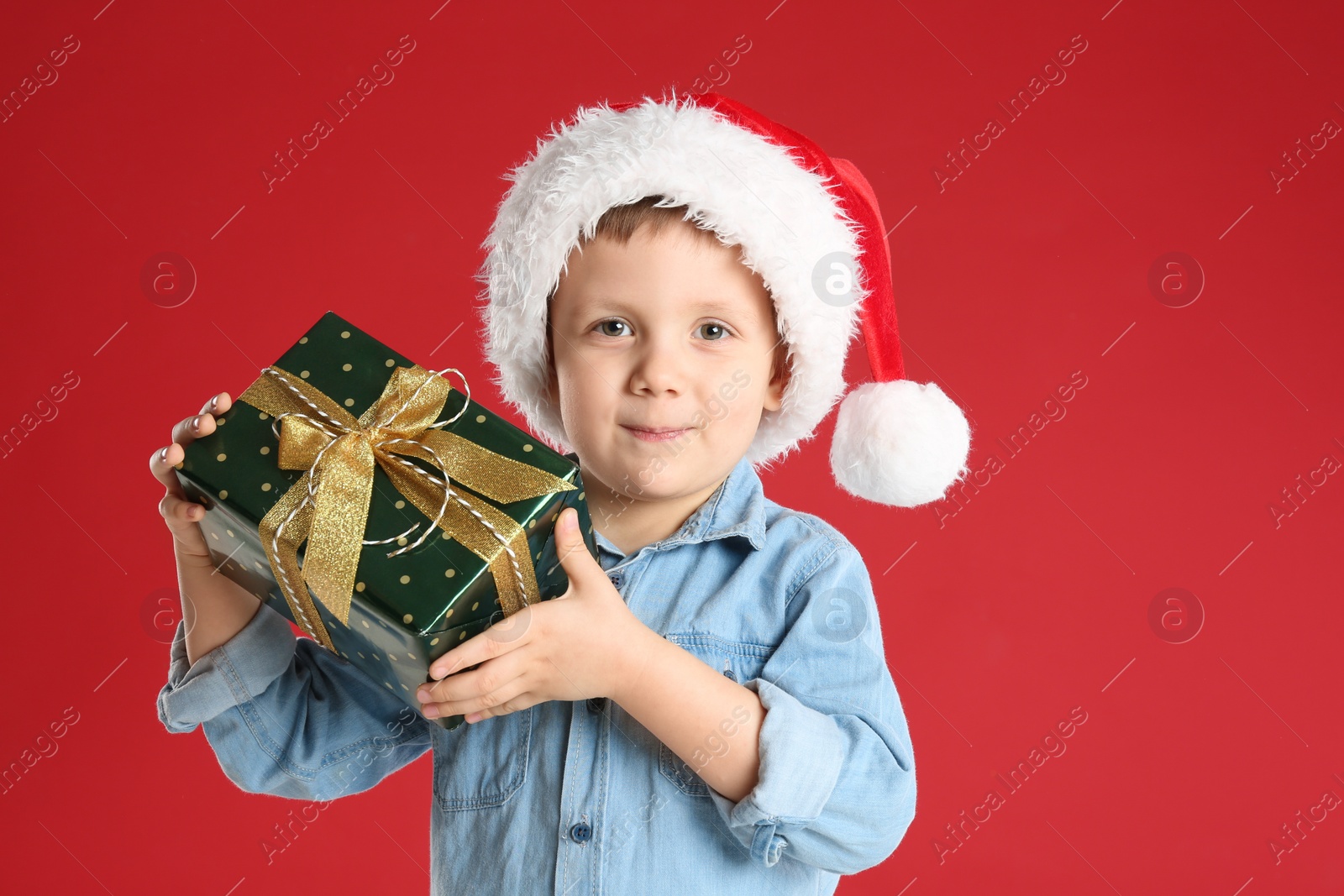 Photo of Cute child in Santa hat with Christmas gift on red background