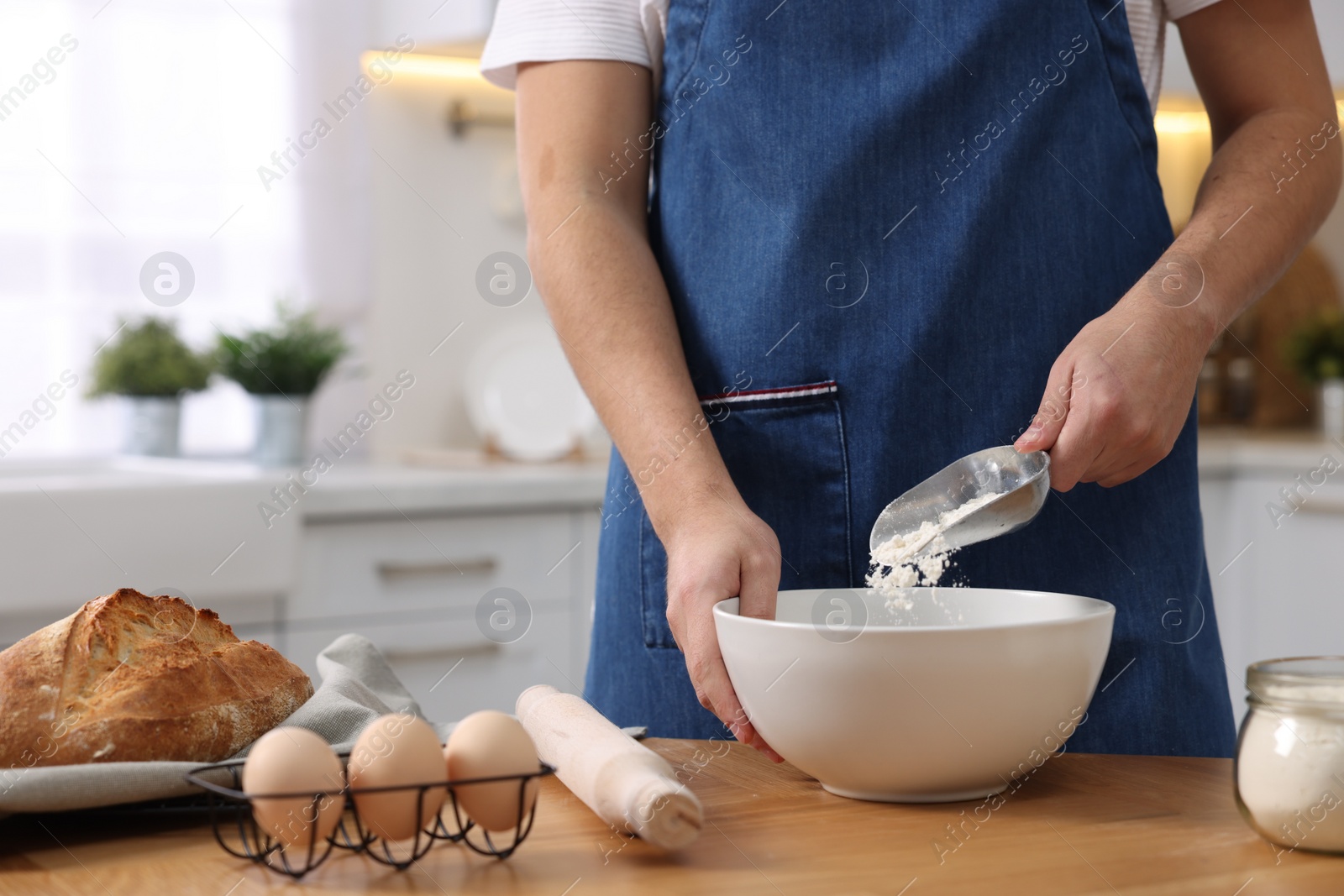 Photo of Making bread. Man putting flour into bowl at wooden table in kitchen, closeup