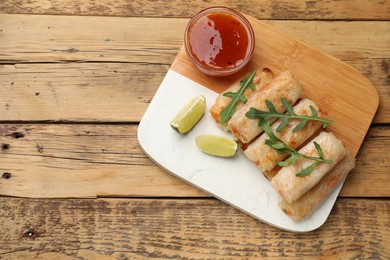 Photo of Tasty fried spring rolls, lime, arugula and sauce on wooden table, top view. Space for text