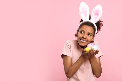 Photo of Happy African American woman in bunny ears headband holding Easter eggs on pink background, space for text