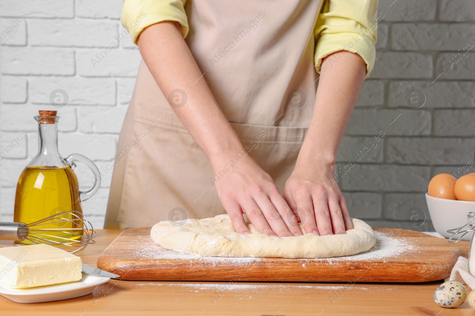 Photo of Woman kneading dough at wooden table near white brick wall, closeup