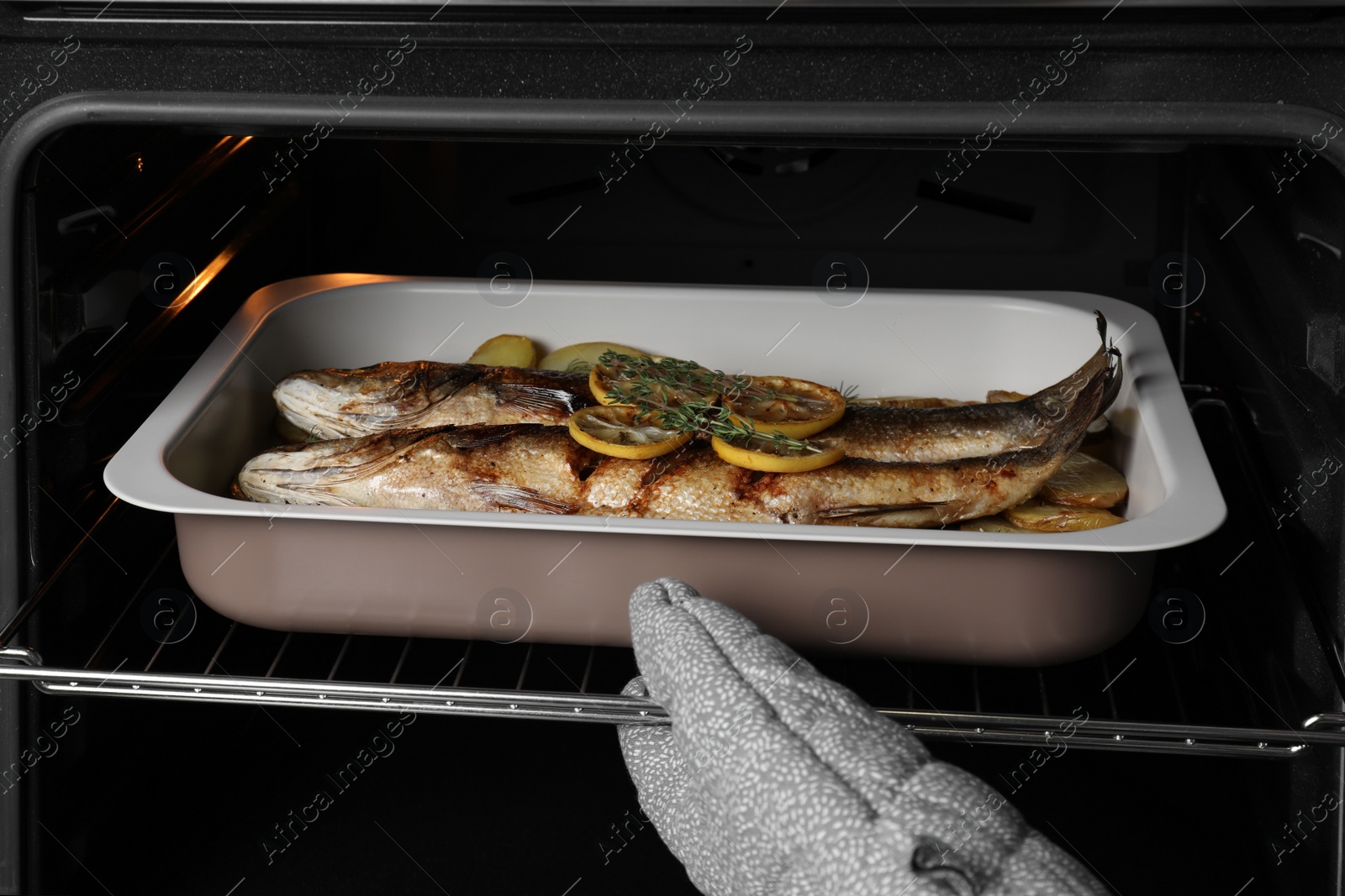 Photo of Woman taking out baking tray with sea bass fish and potatoes from oven