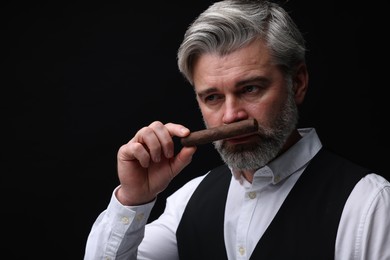 Handsome bearded man smelling cigar on black background. Space for text