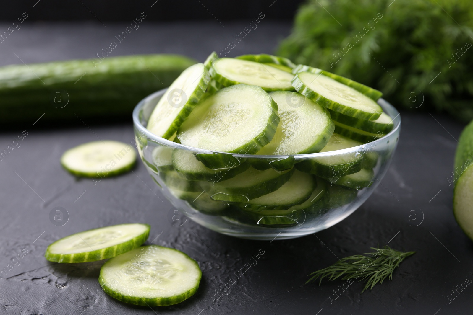 Photo of Cut cucumber in glass bowl on dark gray textured table, closeup