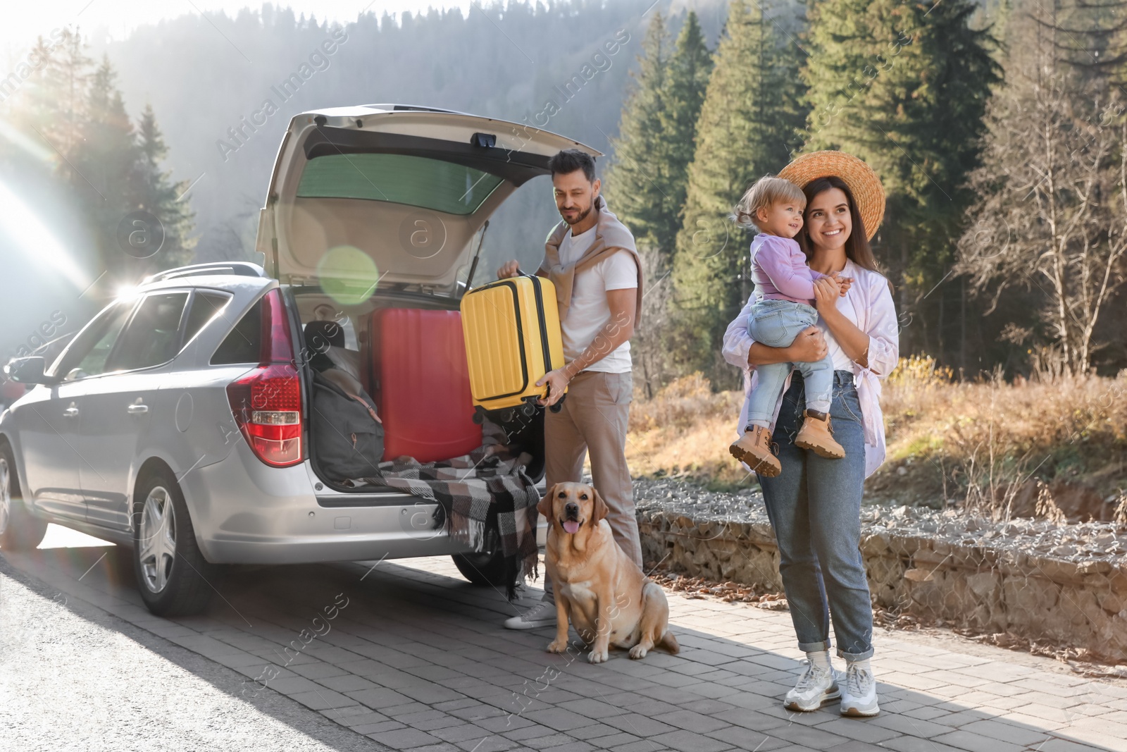 Photo of Mother holding daughter, man and dog near car outdoors. Family traveling with pet