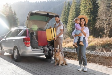 Photo of Mother holding daughter, man and dog near car outdoors. Family traveling with pet