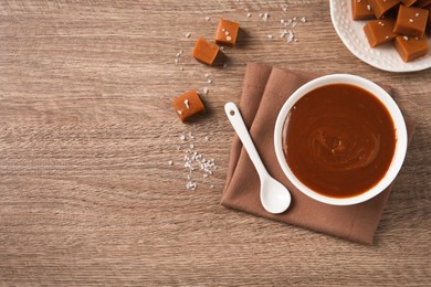 Yummy salted caramel in bowl and candies on wooden table, flat lay. Space for text