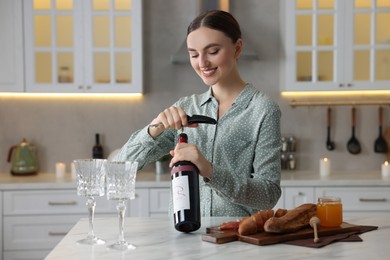Photo of Romantic dinner. Happy woman opening wine bottle with corkscrew at table in kitchen