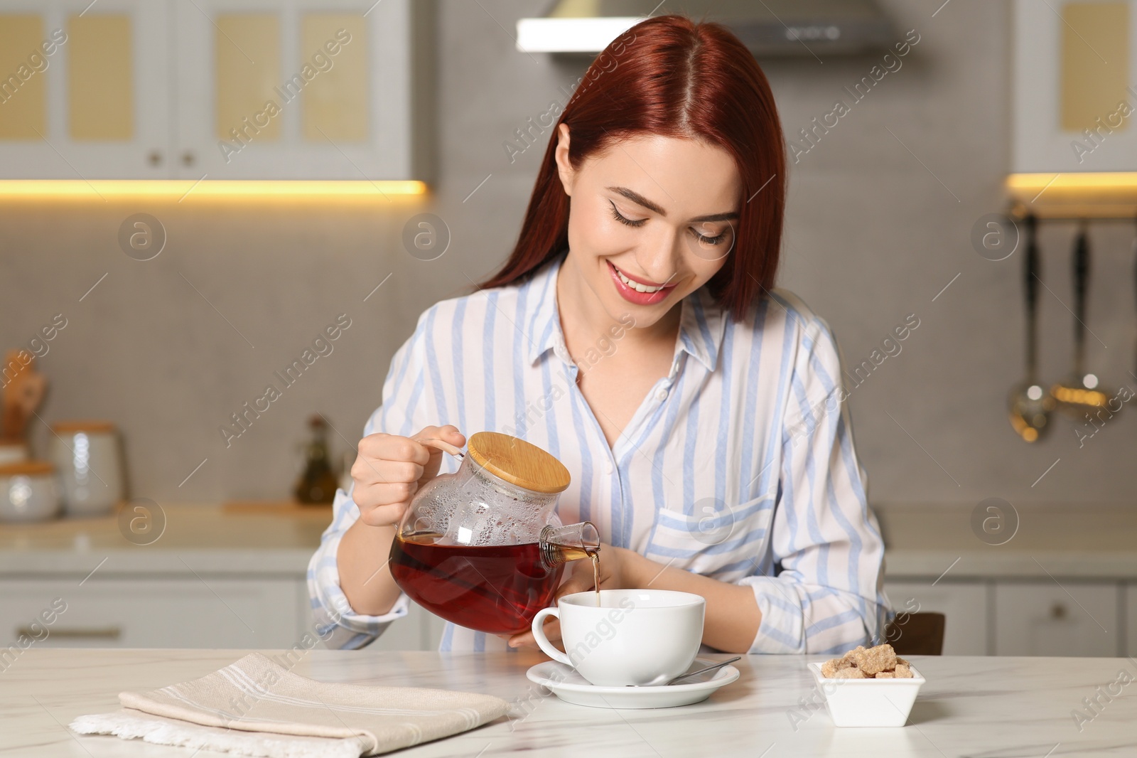 Photo of Happy woman with red dyed hair pouring tea from glass teapot into cup at white table in kitchen