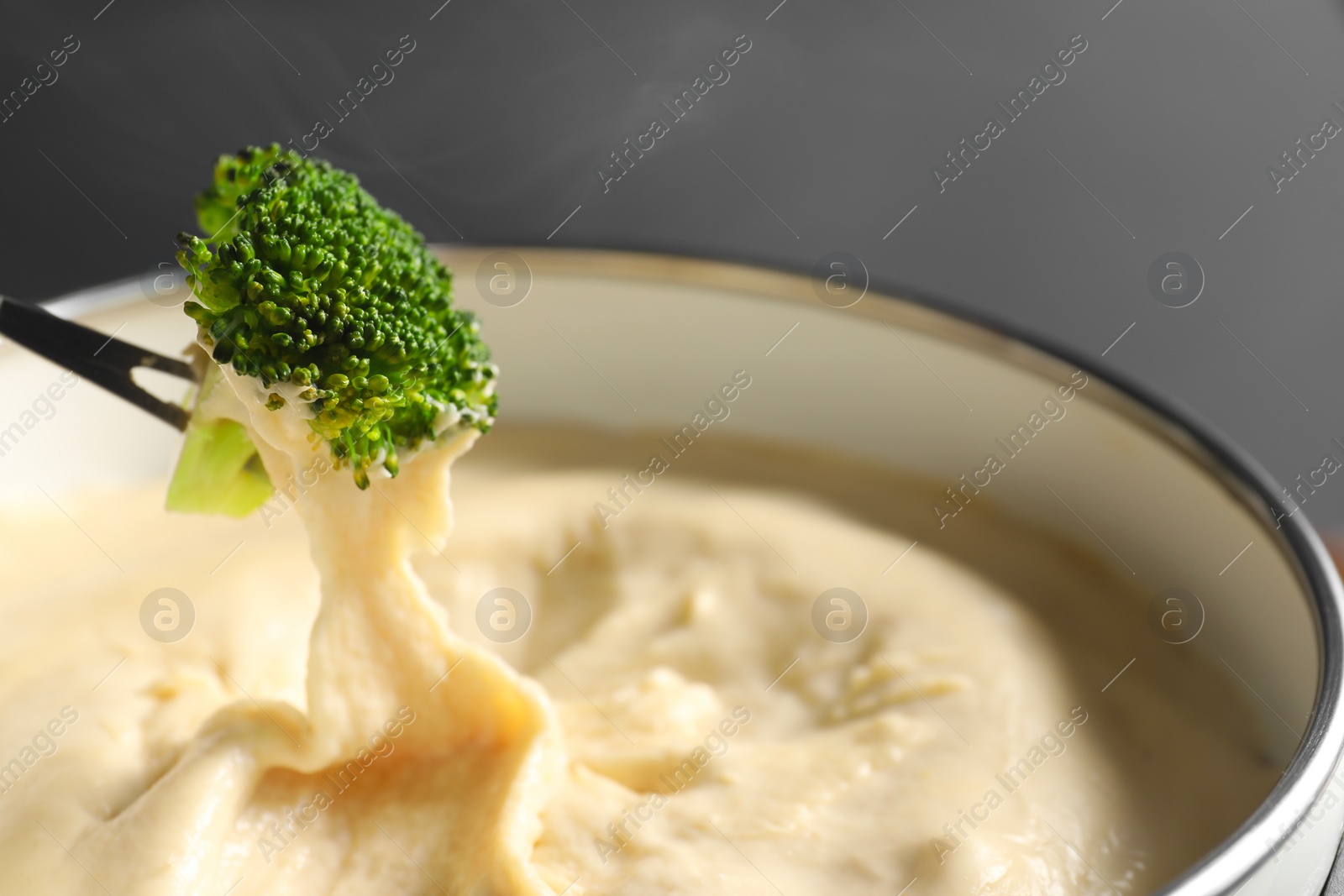 Photo of Dipping piece of broccoli into fondue pot with melted cheese on grey background, closeup