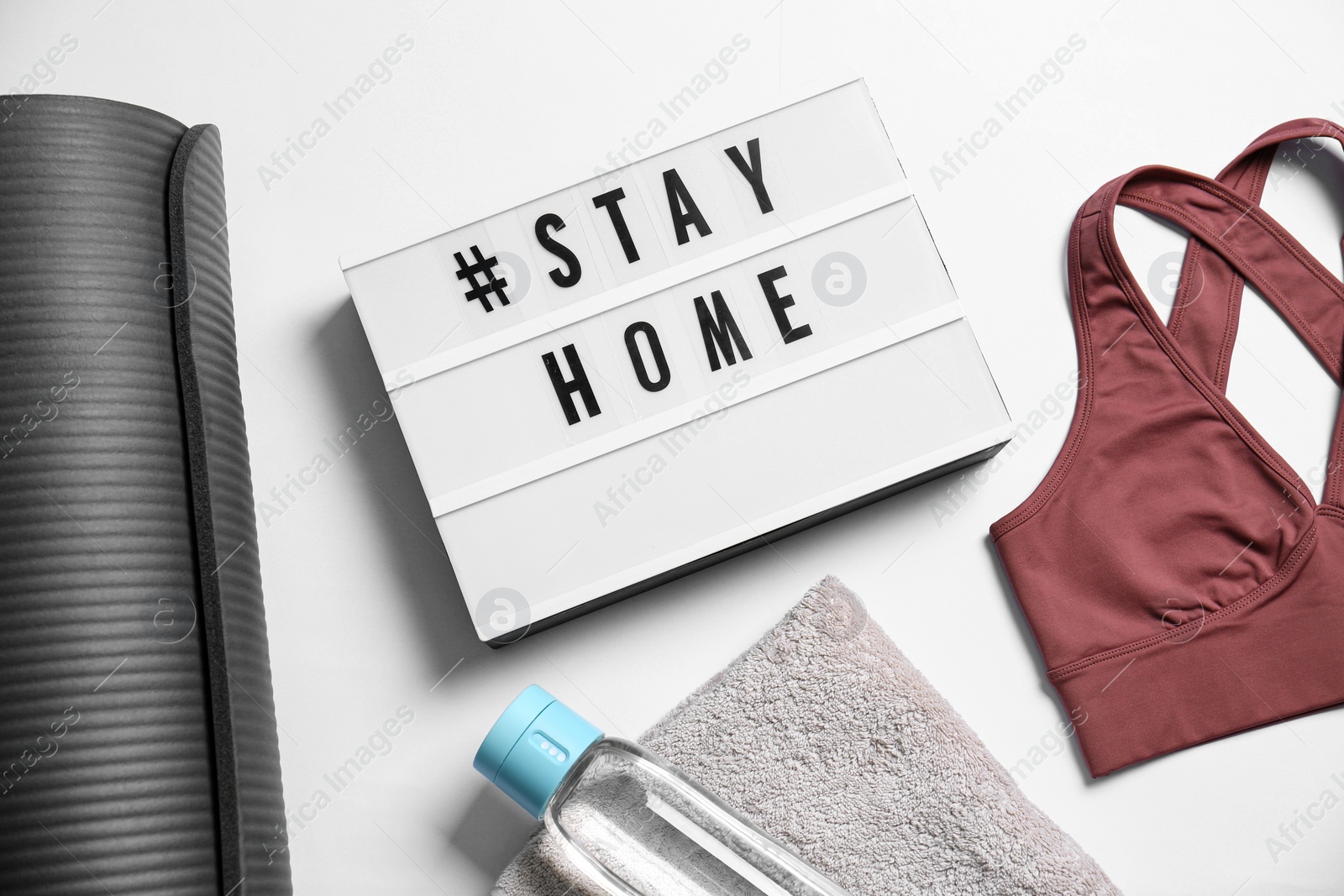 Photo of Stylish sportswear, yoga mat and light box with hashtag Stay at Home on white background, flat lay. Self isolation during COVID‑19 pandemic