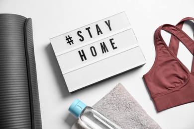 Photo of Stylish sportswear, yoga mat and light box with hashtag Stay at Home on white background, flat lay. Self isolation during COVID‑19 pandemic