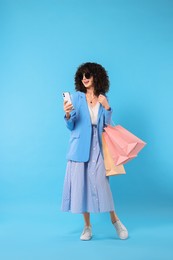 Happy young woman with shopping bags and modern smartphone on light blue background
