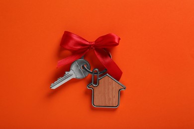 Photo of Key with trinket in shape of house and red bow on orange background, top view. Housewarming party