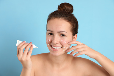 Teen girl with acne problem applying cream on light blue background